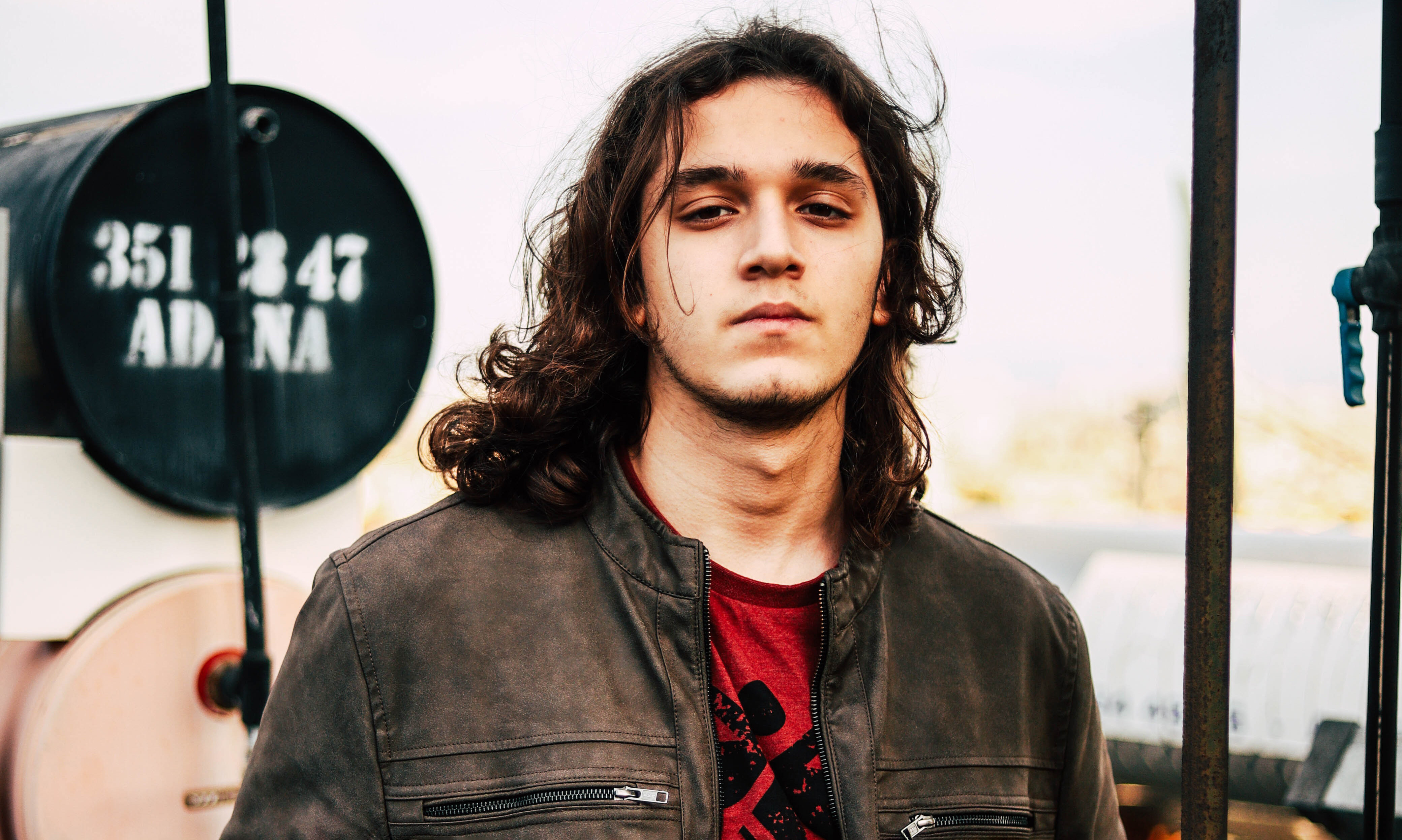 A long-haired man wearing a brown leather jacket and red t-shirt.