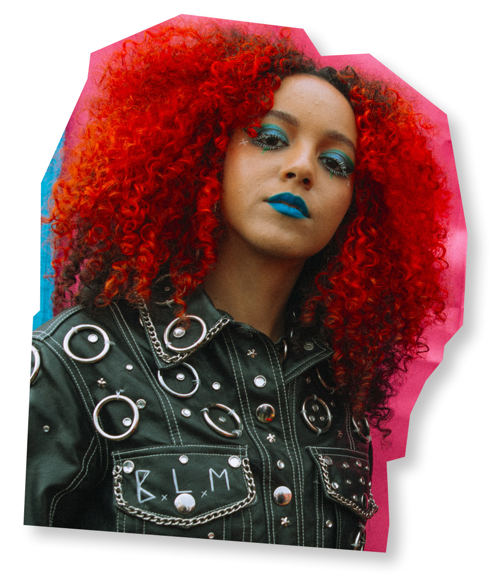 A woman with cyan eyeshadow and lipstick with bright red coiled hair. Her jacket is studded and has B.L.M. written on it.